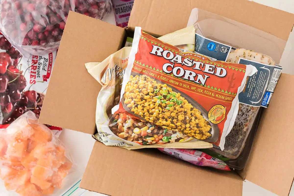 A cardboard box has all four flaps open and is stuffed with frozen corn, chicken and other frozen food getting ready to be packed and shipped in the mail.