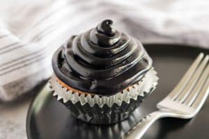 a cupcake sits on a black plate next to a fork with a silver foil cupcake liner topped with jet black icing made from homemade black food coloring mix.