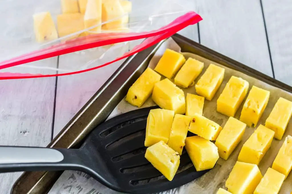 A spatula is being used to transfer frozen pineapple from a sheet pan into an airtight zip lock bag for long term freezer storage.